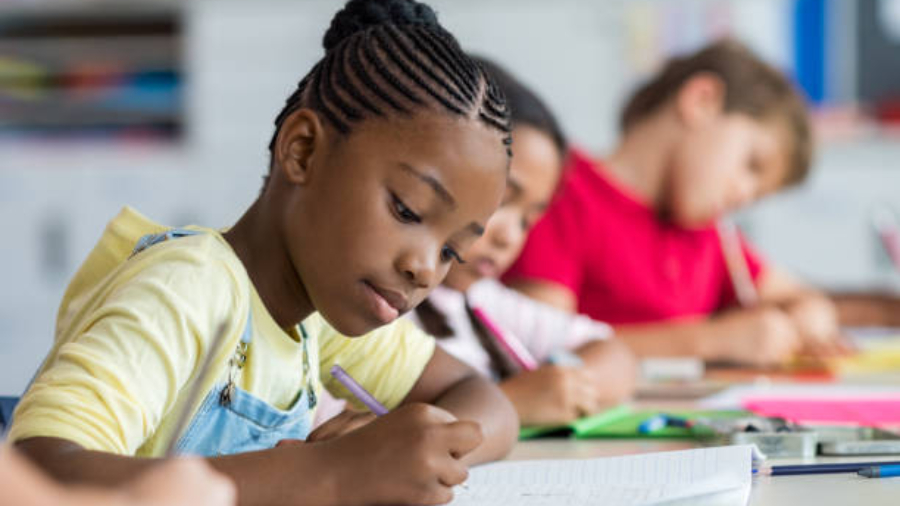 Cute pupil writing at desk in classroom at the elementary school. Student girl doing test in primary school. Children writing notes in classroom. African schoolgirl writing on notebook during the lesson.